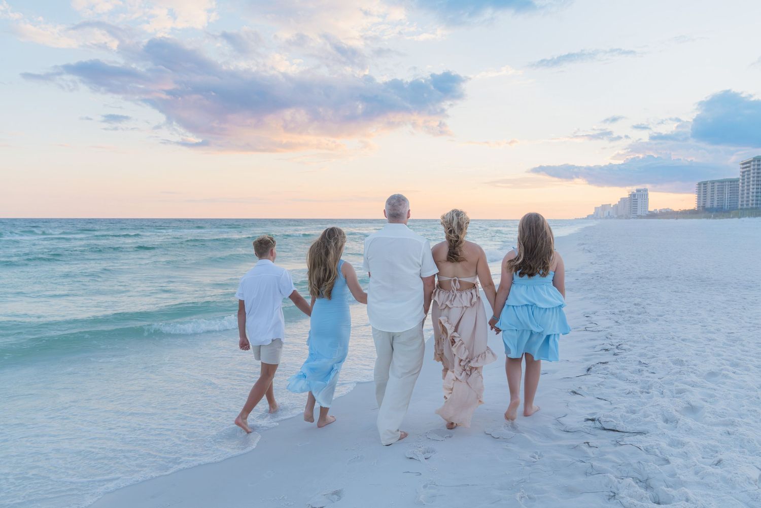 Family of five walking hand-in-hand on a white sand beach at sunset, dressed in coordinating white and blue outfits.
