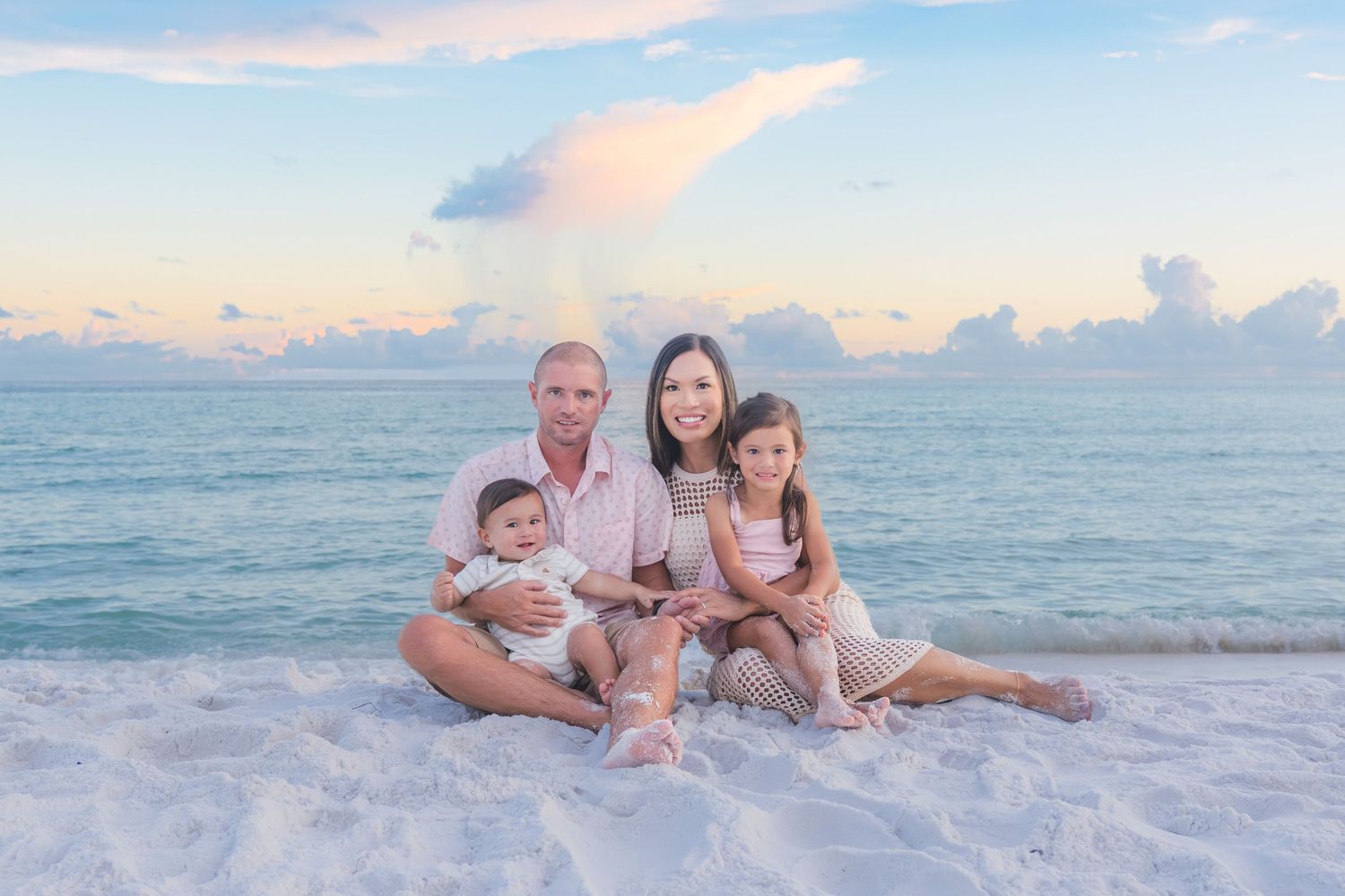 Family of four sitting on white sand beach at sunset with calm turquoise ocean and pastel sky behind them.
