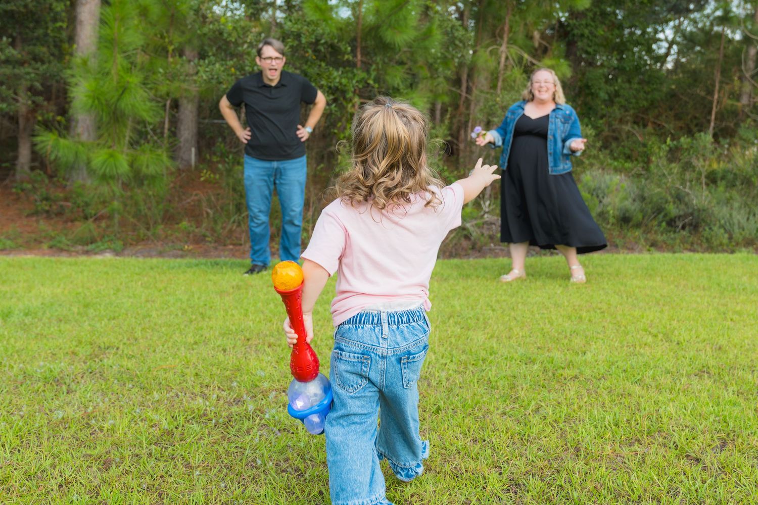 A young girl holds a toy bat while playing with two adults on a green lawn surrounded by pine trees.