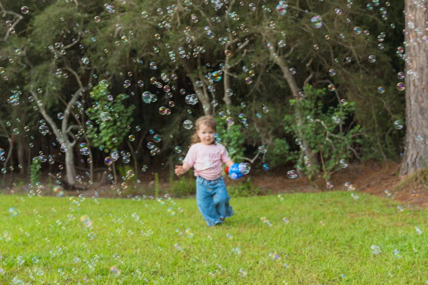 Young child running through green grass park chasing colorful soap bubbles floating in the air on a sunny day.