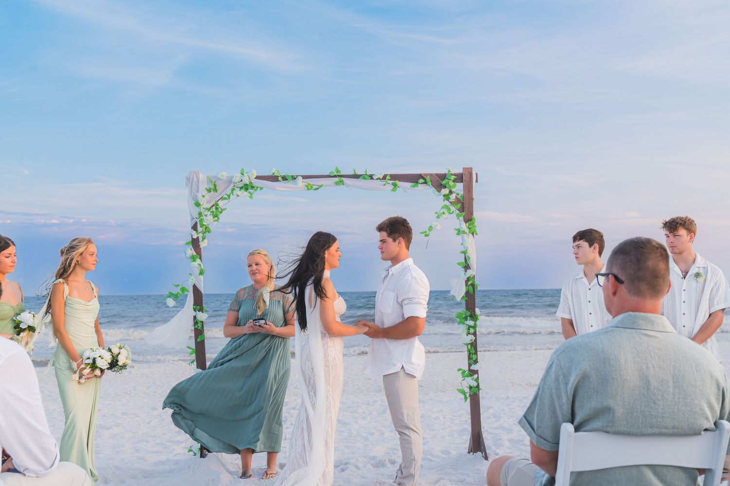 Beach wedding ceremony with couple exchanging vows under floral arch, bridal party in sage green, ocean backdrop at sunset.