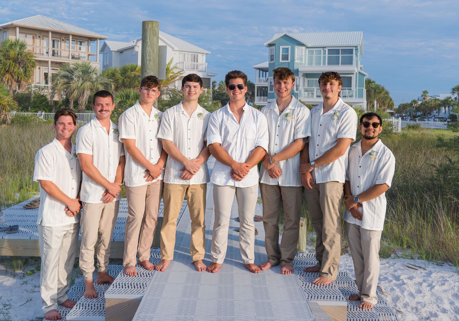Group of eight young men in white shirts posing on a beach boardwalk for a wedding photo.