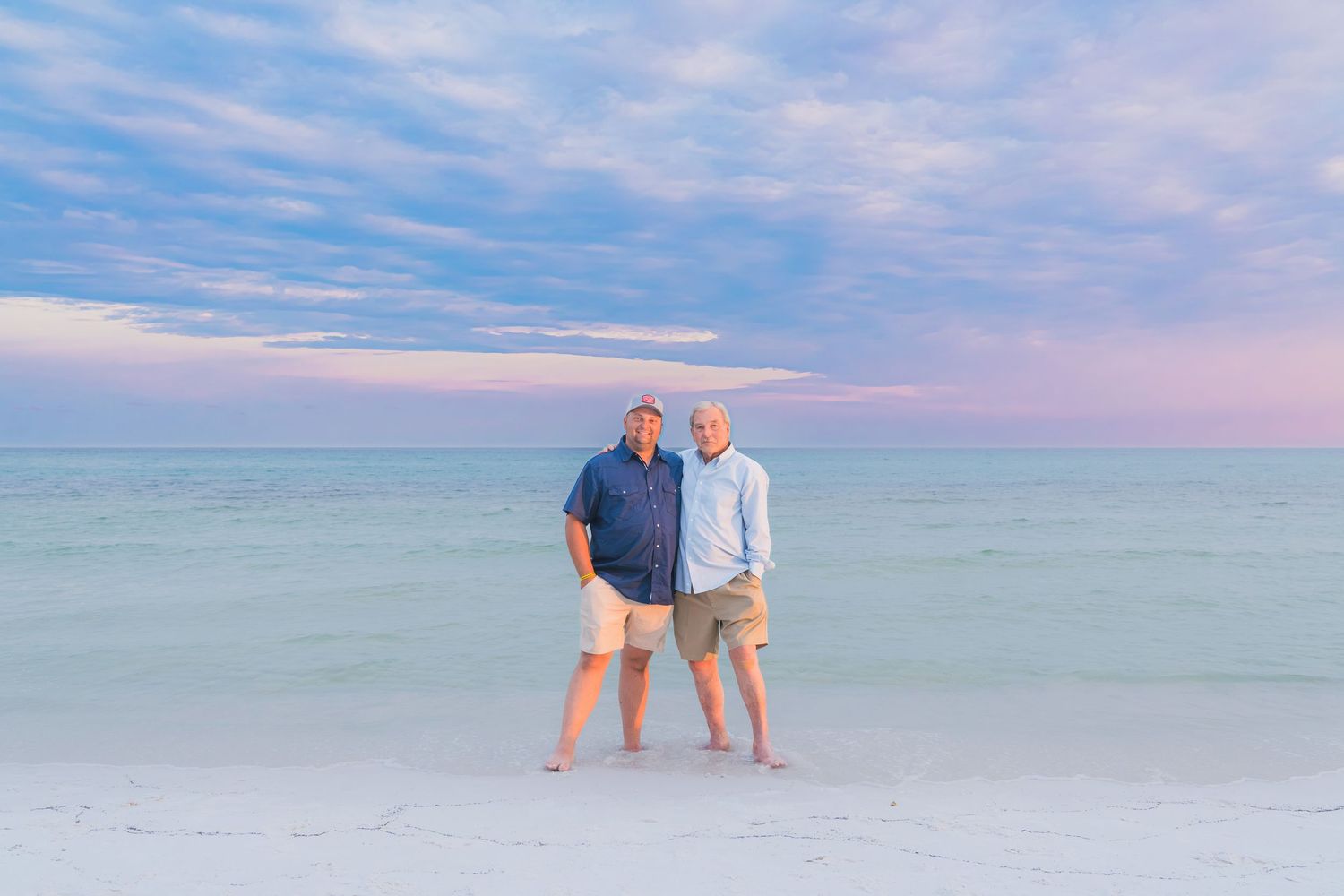 Two men smile together on a white sand beach with pastel sunset sky and calm ocean behind them.