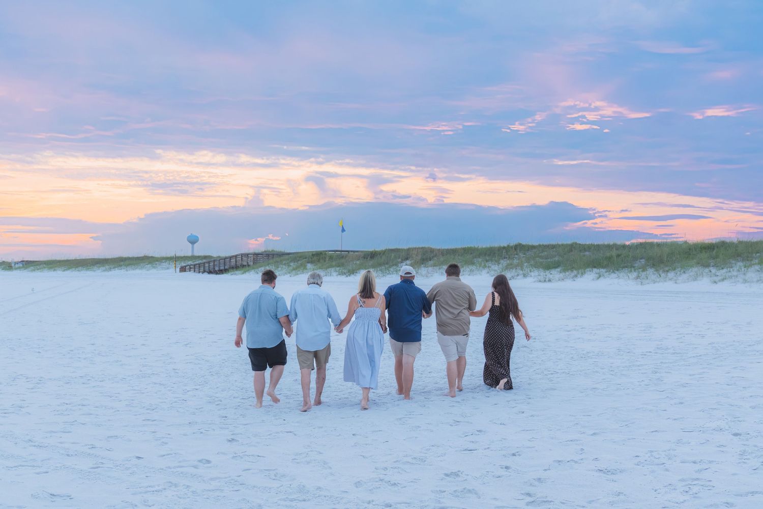 Six people walk hand-in-hand on a white sand beach at sunset with colorful pink and blue skies.