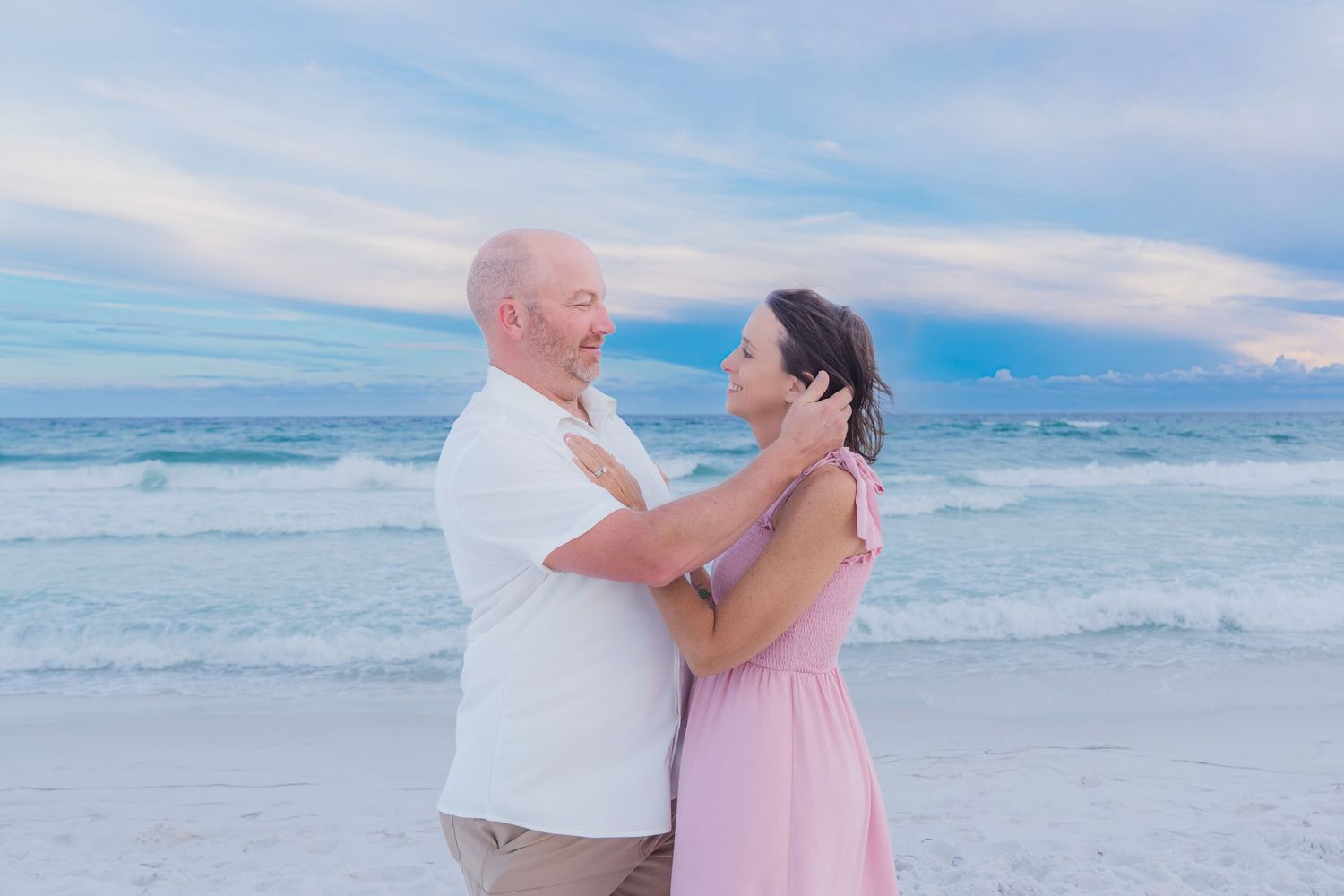 Couple sharing a romantic moment on a sandy beach at sunset, with turquoise ocean waves and pastel sky behind them.