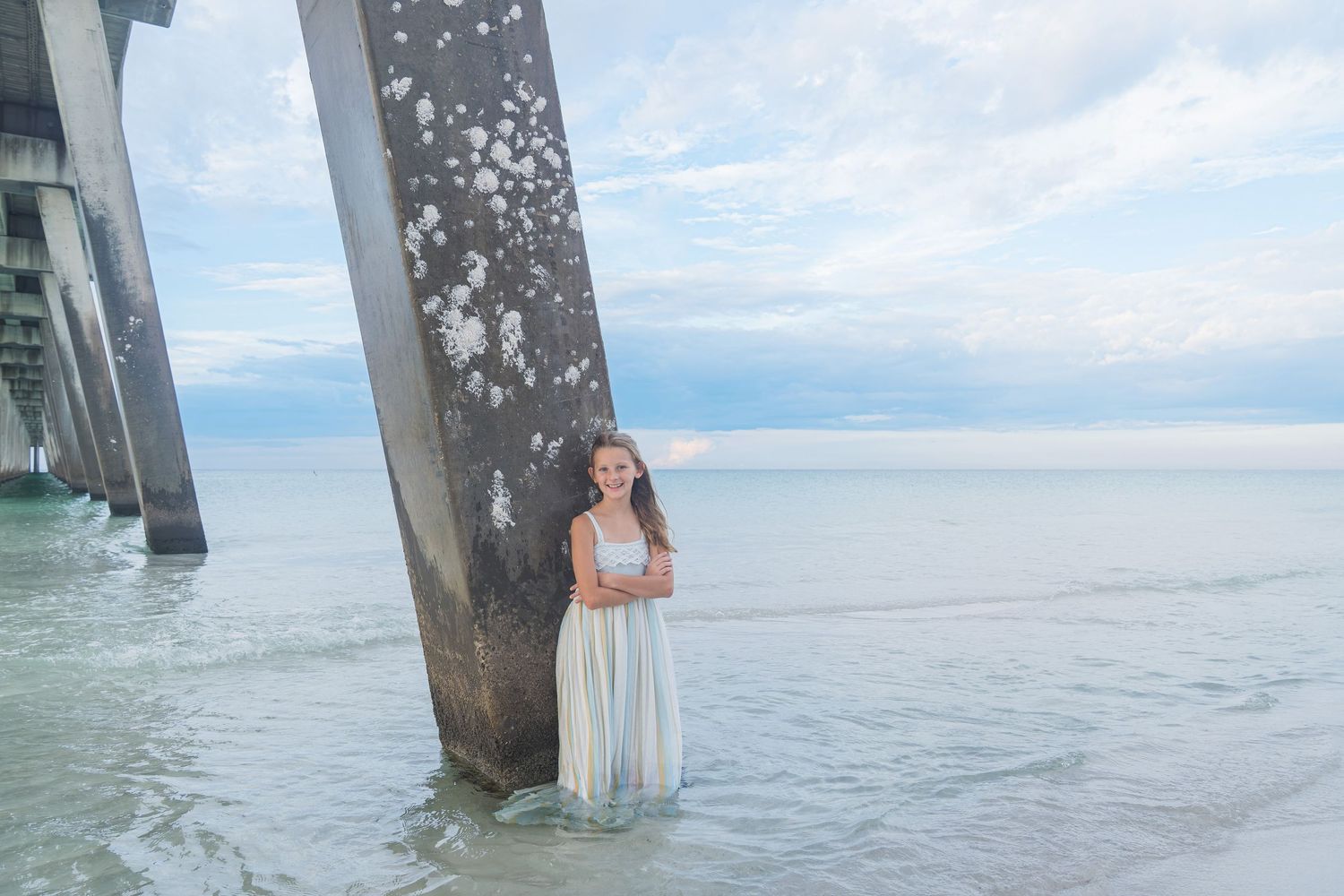 Young girl in white dress standing by pier pillar in ocean water.