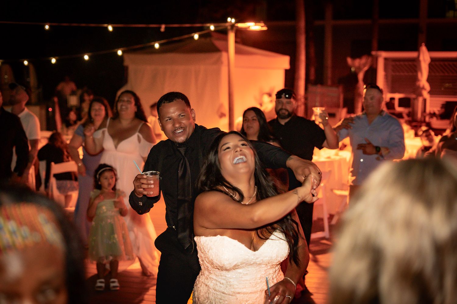 Bride and groom laughing and dancing joyfully at outdoor wedding reception with warm string lights at night.