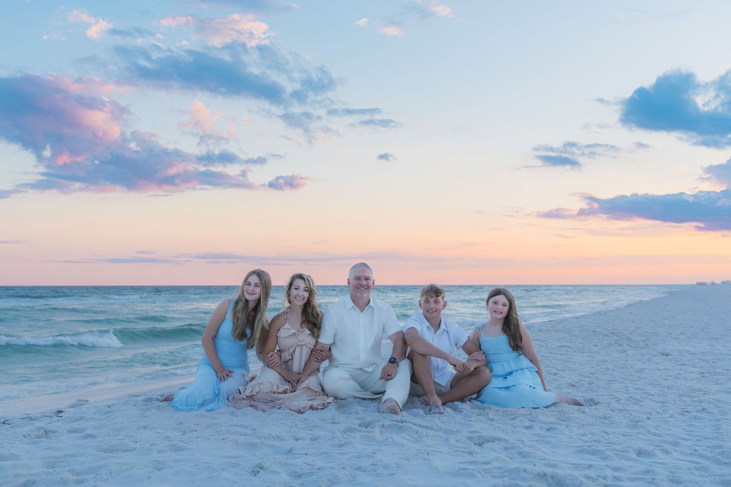 Family of five posing on sandy beach at sunset with pastel pink and blue sky over calm ocean waters.