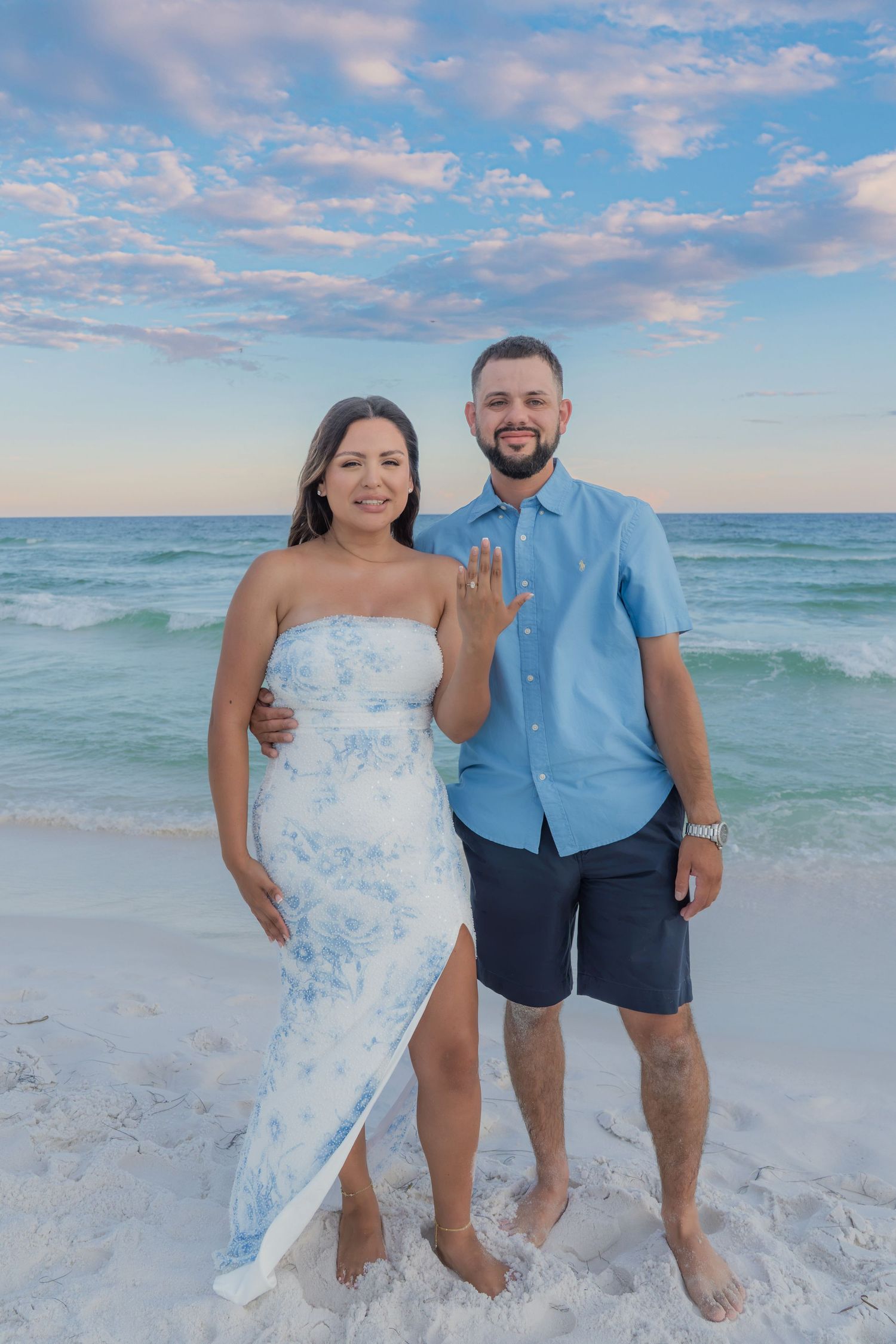 Couple poses on white sand beach at sunset, woman in floral dress showing engagement ring, man in blue shirt and shorts.