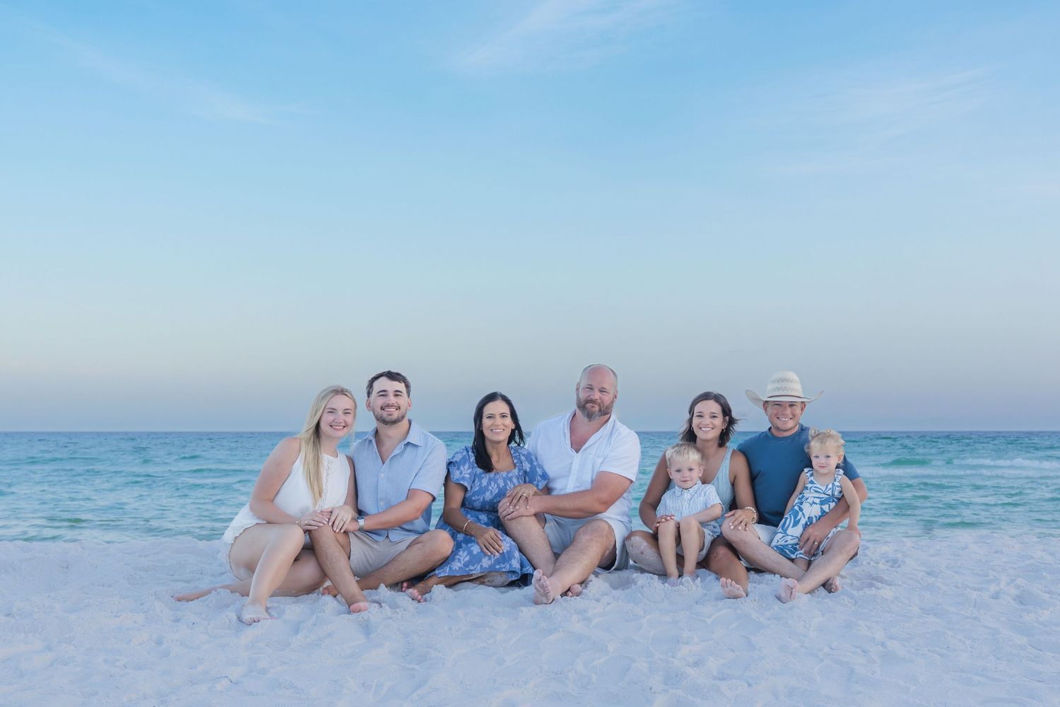 Family of seven posing together on a white sandy beach at sunset with calm turquoise ocean waters in the background.