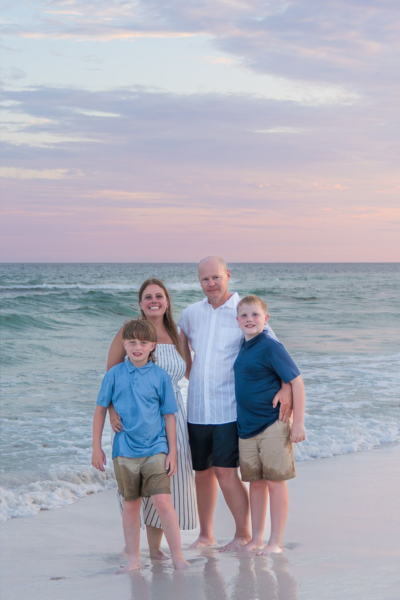 Happy family of four posing on a sandy beach with pastel sunset sky and ocean waves.