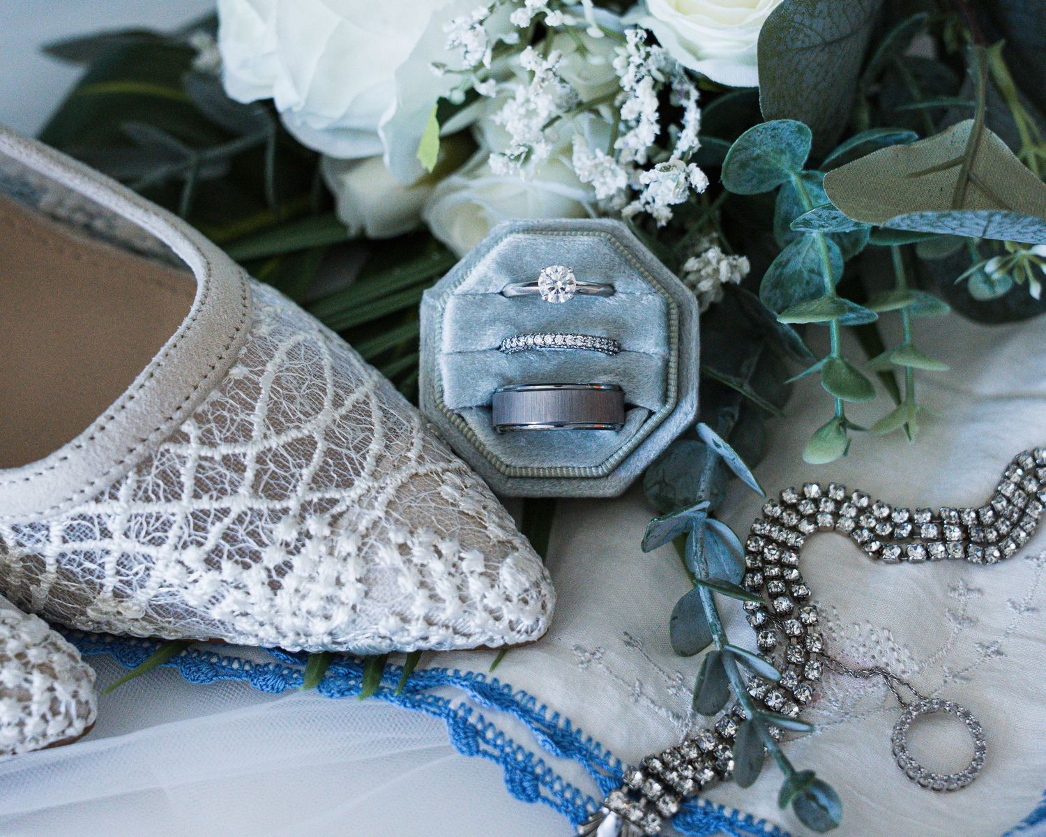 Wedding flat lay featuring lace bridal shoes, diamond rings in a velvet box, and elegant jewelry on white florals.