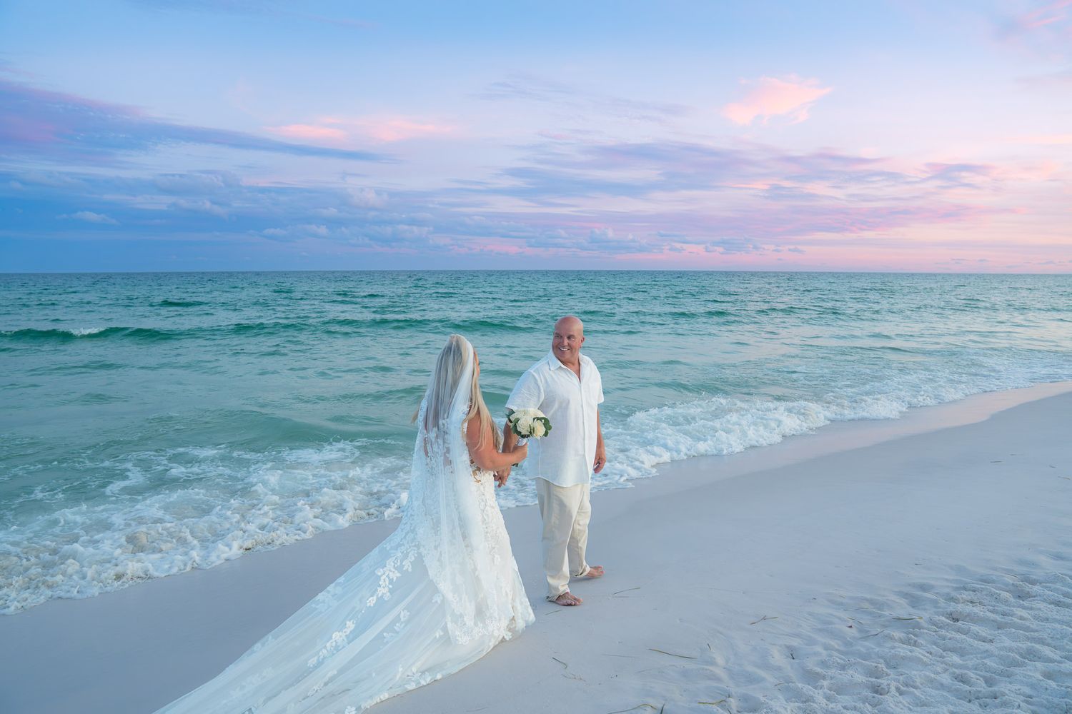 Bride and groom holding hands on a white sand beach at sunset with turquoise ocean waves and pastel sky.