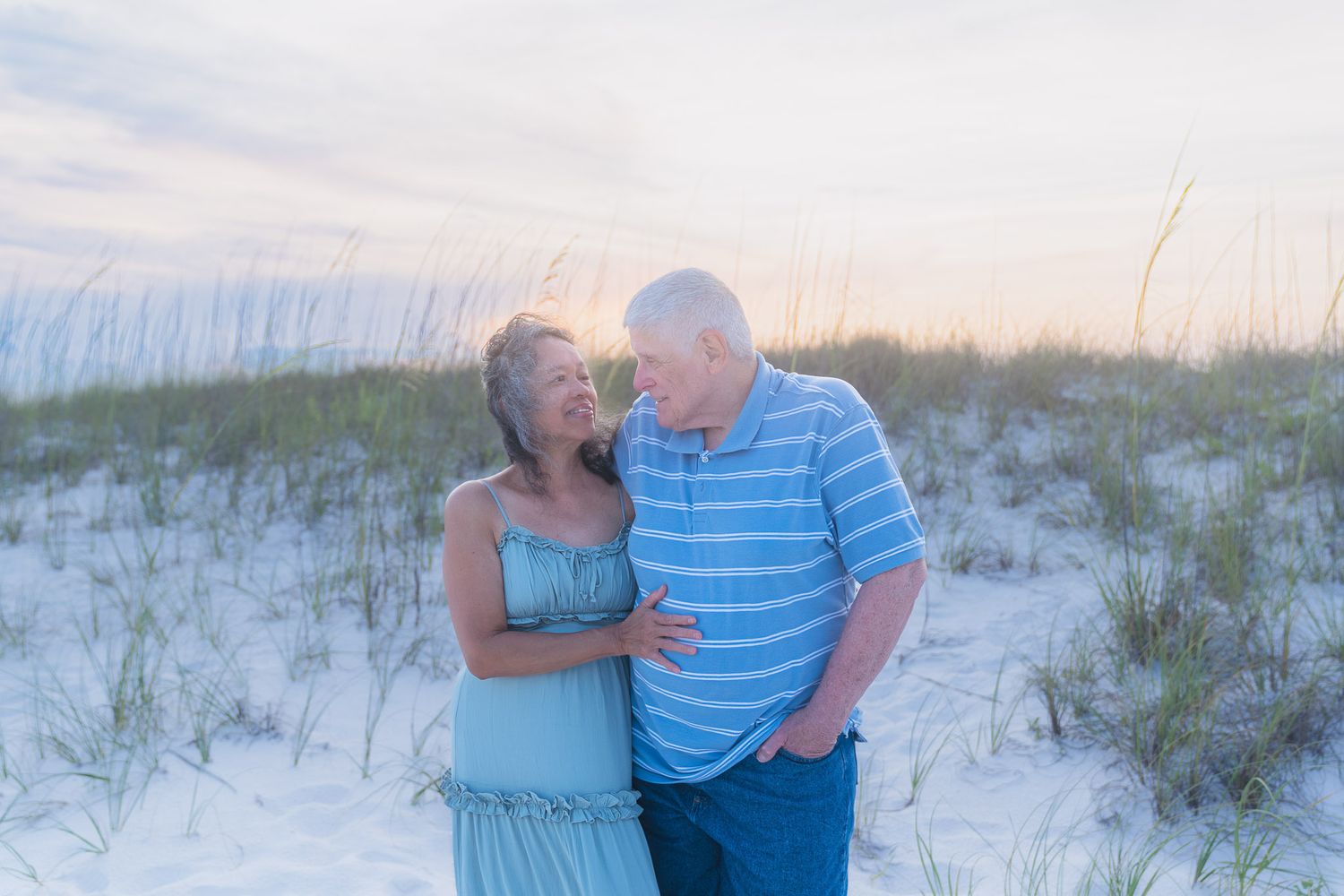 Couple embracing on white sand beach at sunset near sea oats and sand dunes.