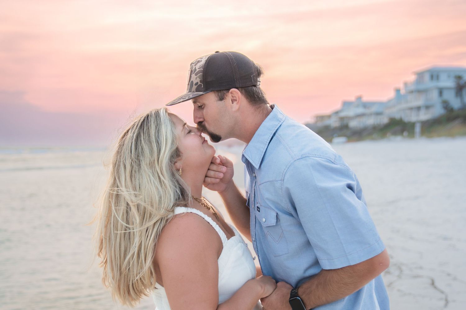 Couple sharing a romantic kiss on the beach at sunset with a pink and purple sky backdrop.