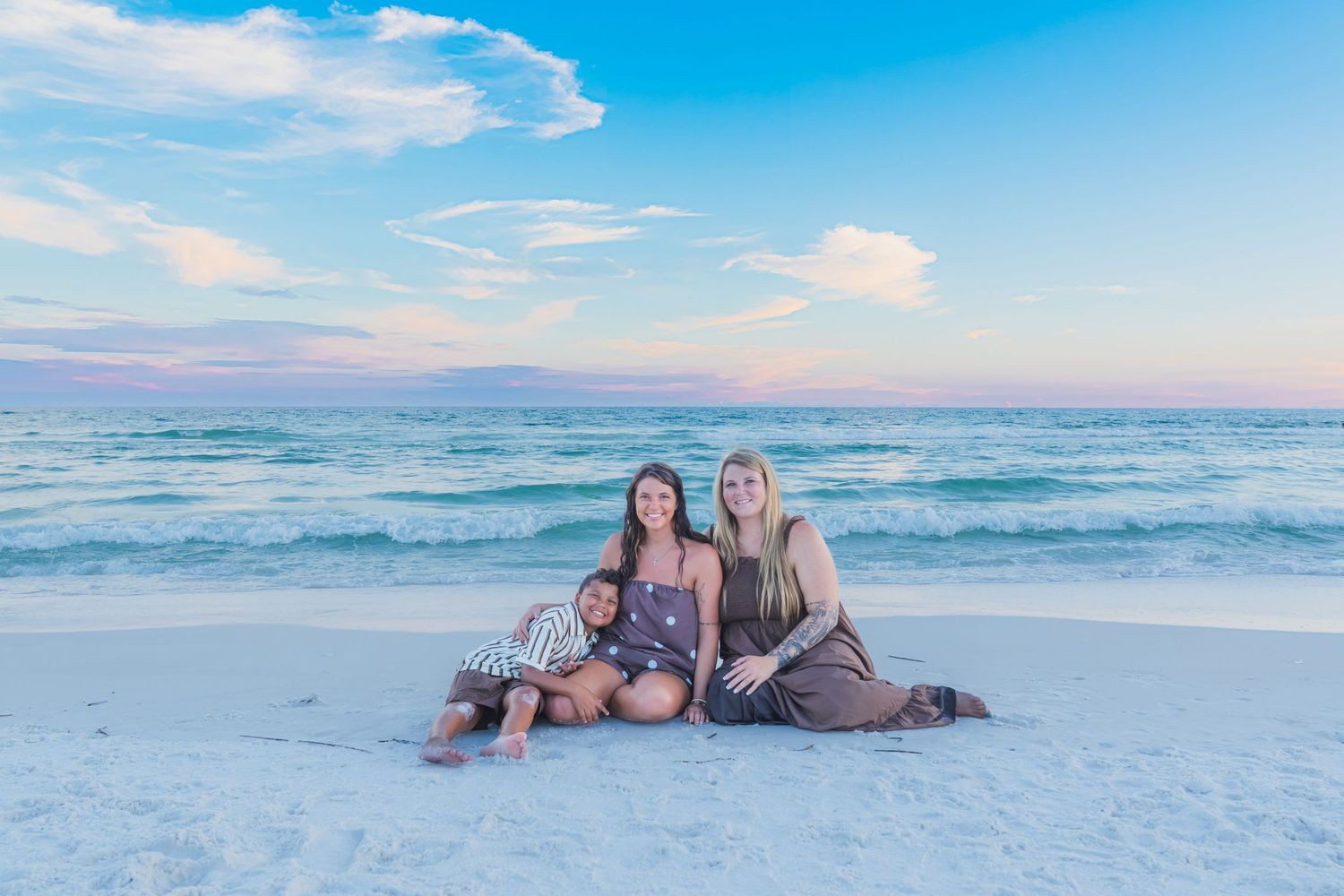 Family of three posing on white sand beach at sunset with turquoise ocean waves in the background.
