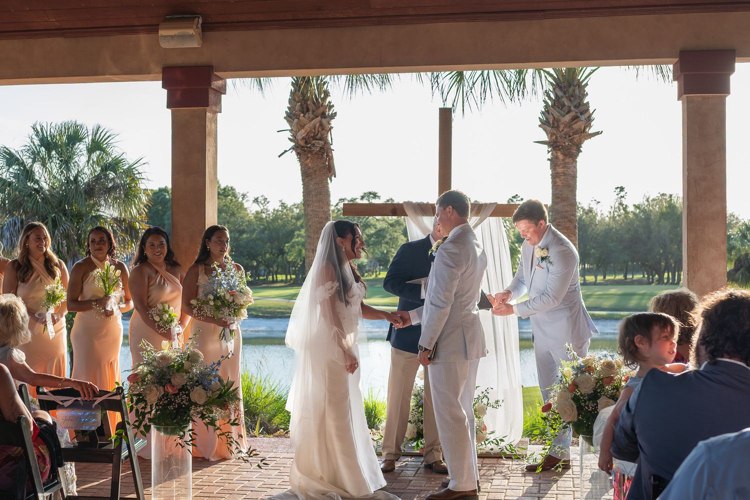 Bride and groom exchange vows at outdoor wedding ceremony with bridesmaids and water view backdrop.