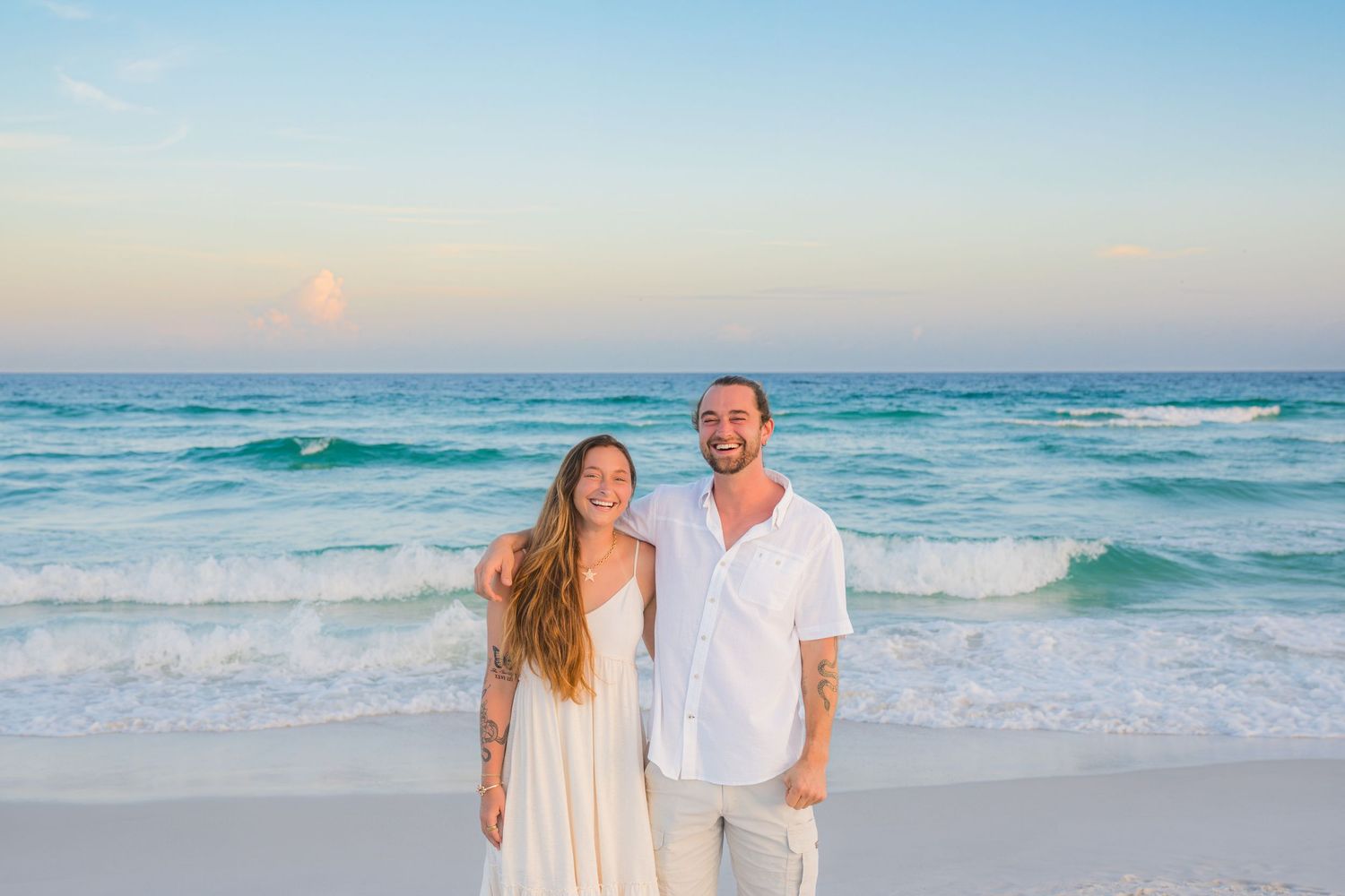 Smiling couple in white outfits posing on a white sand beach with turquoise waves at sunset.