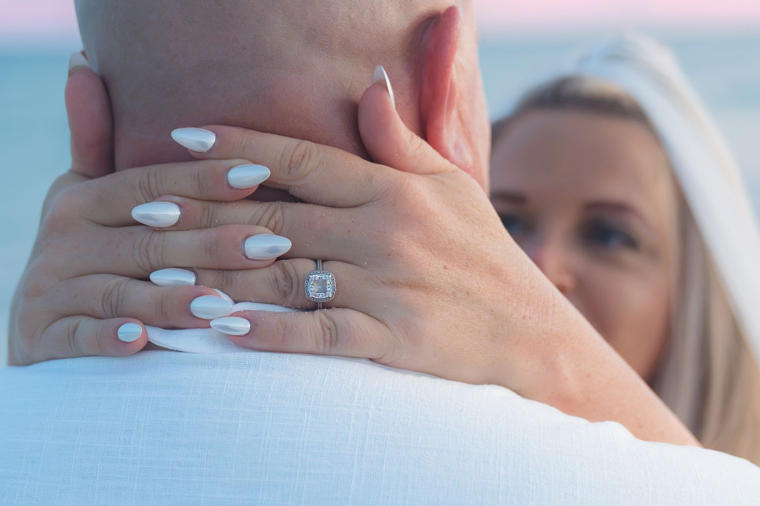 Woman showing diamond engagement ring on her hand while embracing a man at a beach during sunset.