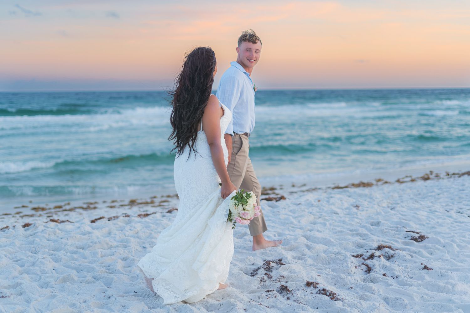 Bride and groom walking hand in hand on a white sandy beach at sunset during their beach wedding.