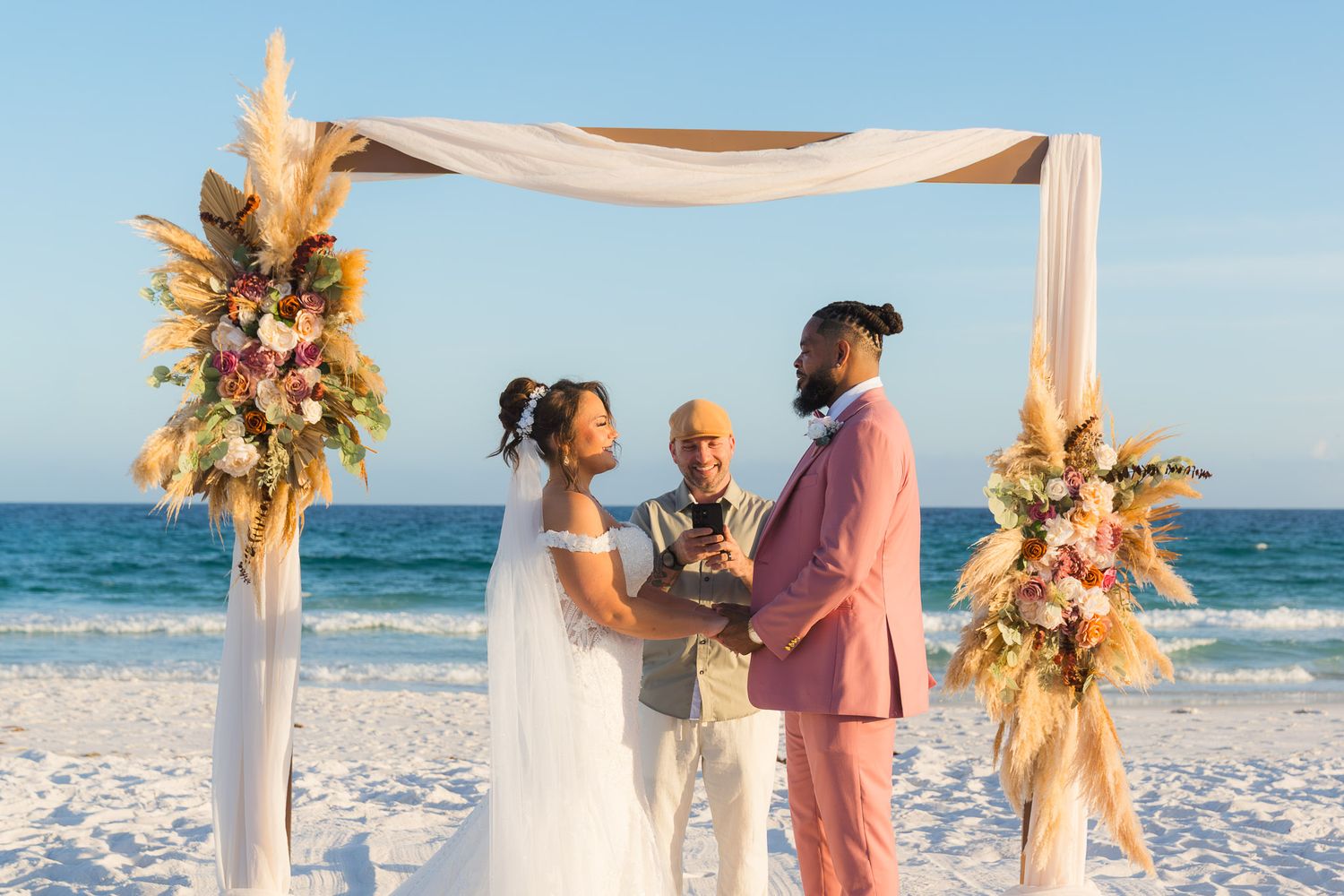 Couple exchanges vows at a beach wedding ceremony under a decorated arch with floral arrangements by the ocean.