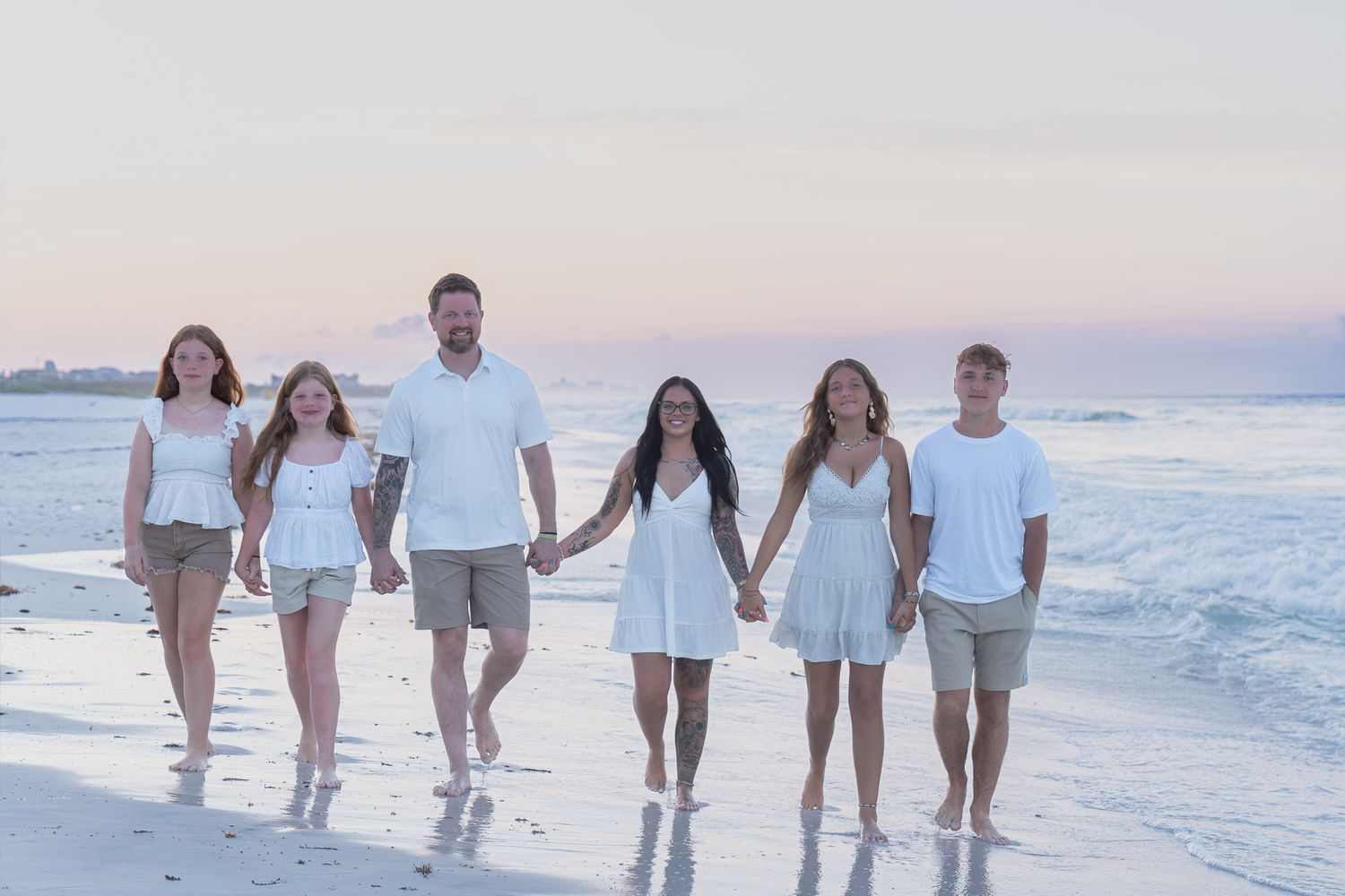Family of six walks hand-in-hand on a sandy beach at sunset, wearing white and neutral outfits.