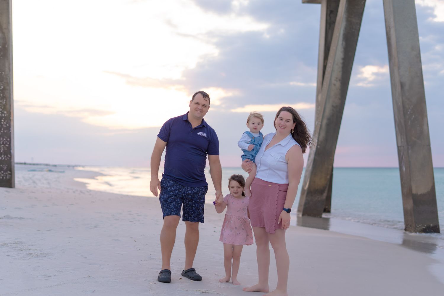 Family of four poses on a white sand beach at sunset near a pier with turquoise water in the background.