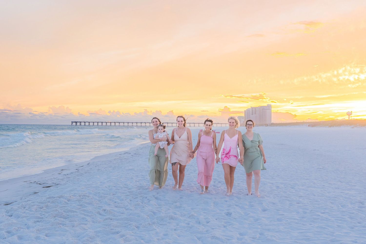 Group of five women in colorful dresses walking on beach at golden sunset with pier in background.