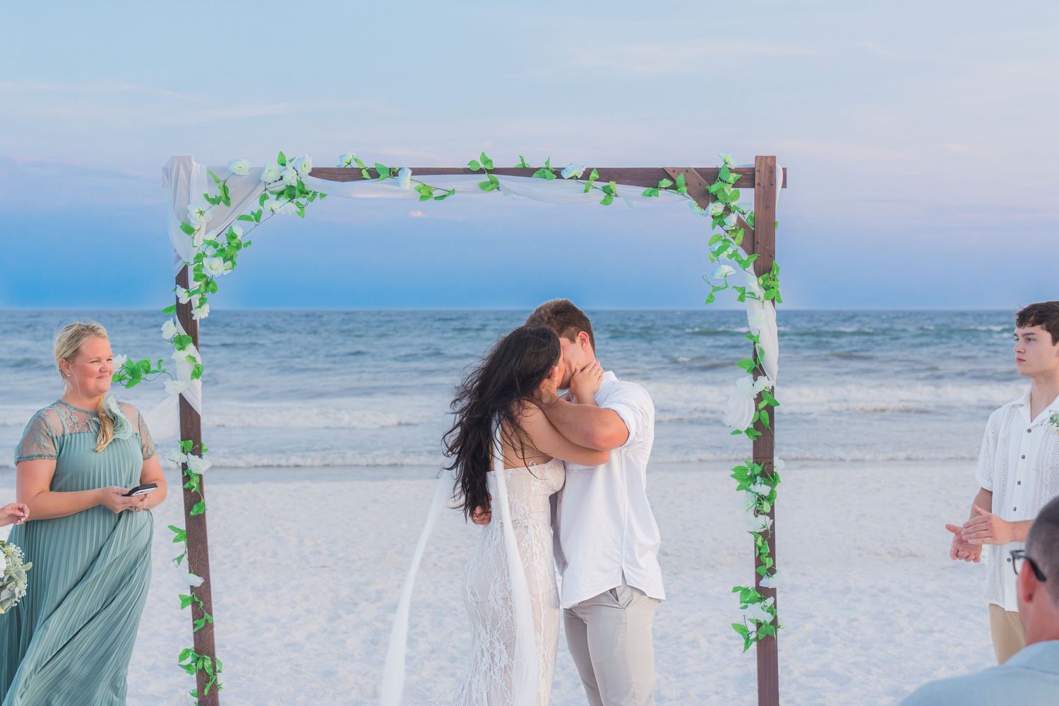 Couple sharing first kiss under floral arch at romantic beach wedding ceremony at sunset.