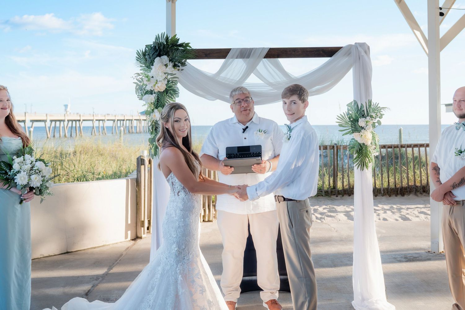 Couple exchanges vows at a beachside wedding ceremony under a white draped arch with floral arrangements.