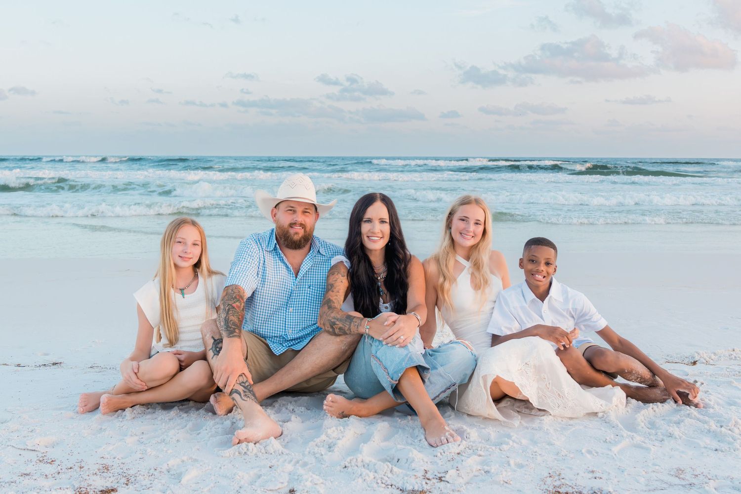 Family of five sitting together on a white sand beach, smiling, with ocean waves and pastel sky in the background.