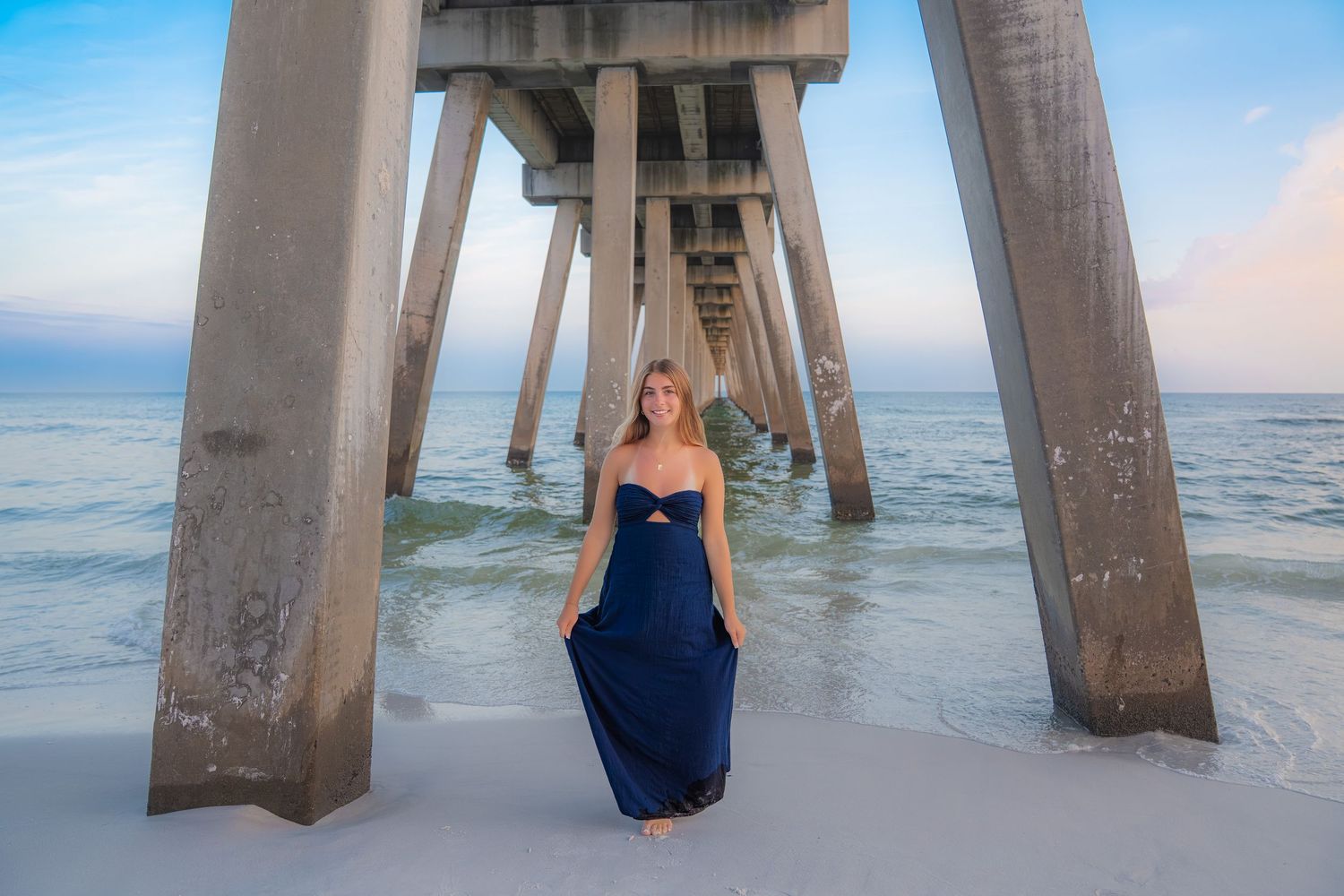 Woman in navy blue maxi dress posing under a beach pier with ocean waves and pastel sky backdrop.