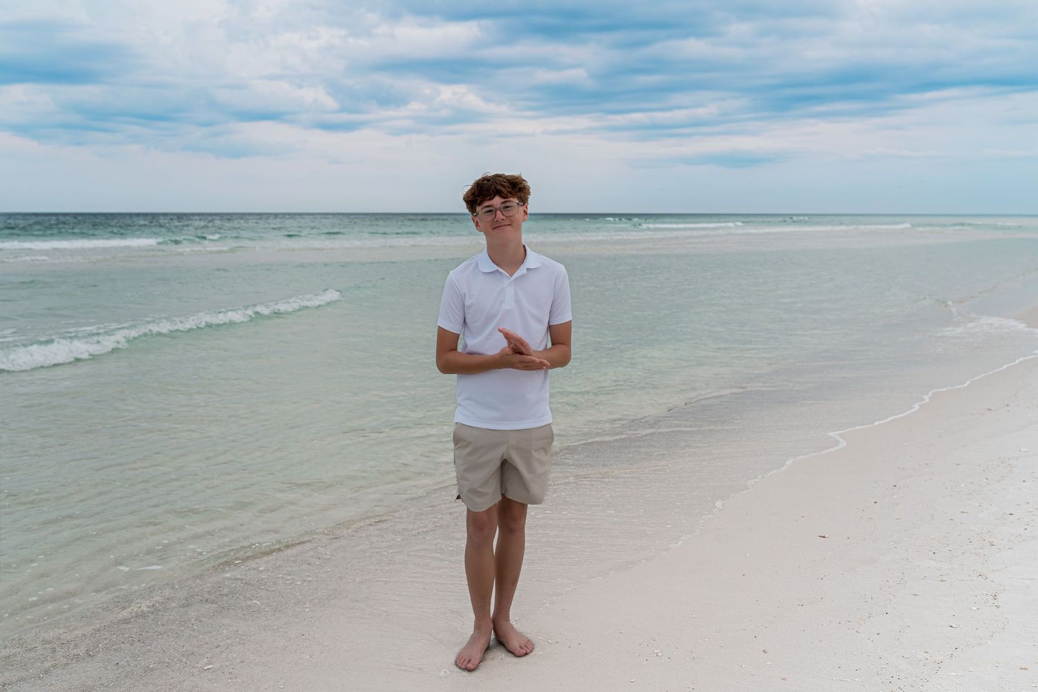 Young man in white polo and khaki shorts standing barefoot on a white sandy beach with calm turquoise water.
