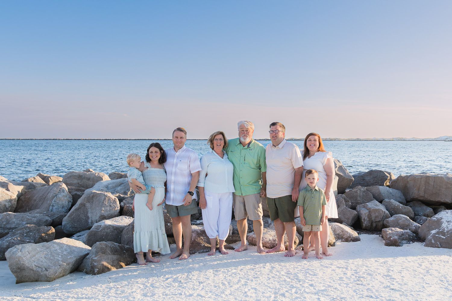 Multi-generational family posing together on a white sand beach with rocky shoreline and calm blue water at sunset.