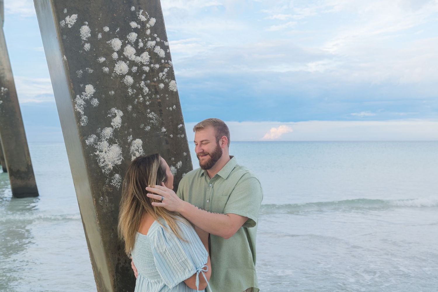 Couple sharing a romantic moment by a beach pier piling with ocean waves in the background at dusk.