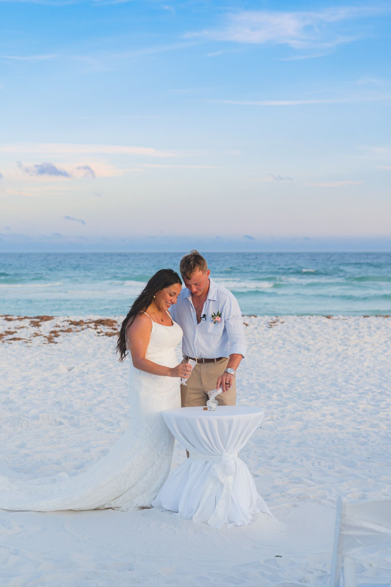 Bride and groom signing documents at a white cocktail table on a sandy beach at sunset.