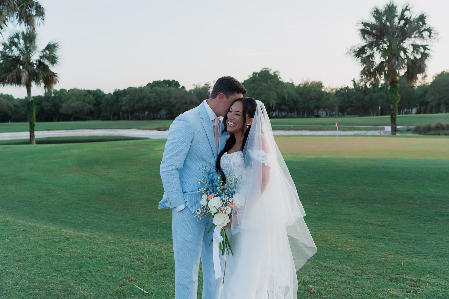 Newlyweds share romantic moment on golf course at sunset, bride holding pastel bouquet with veil flowing.