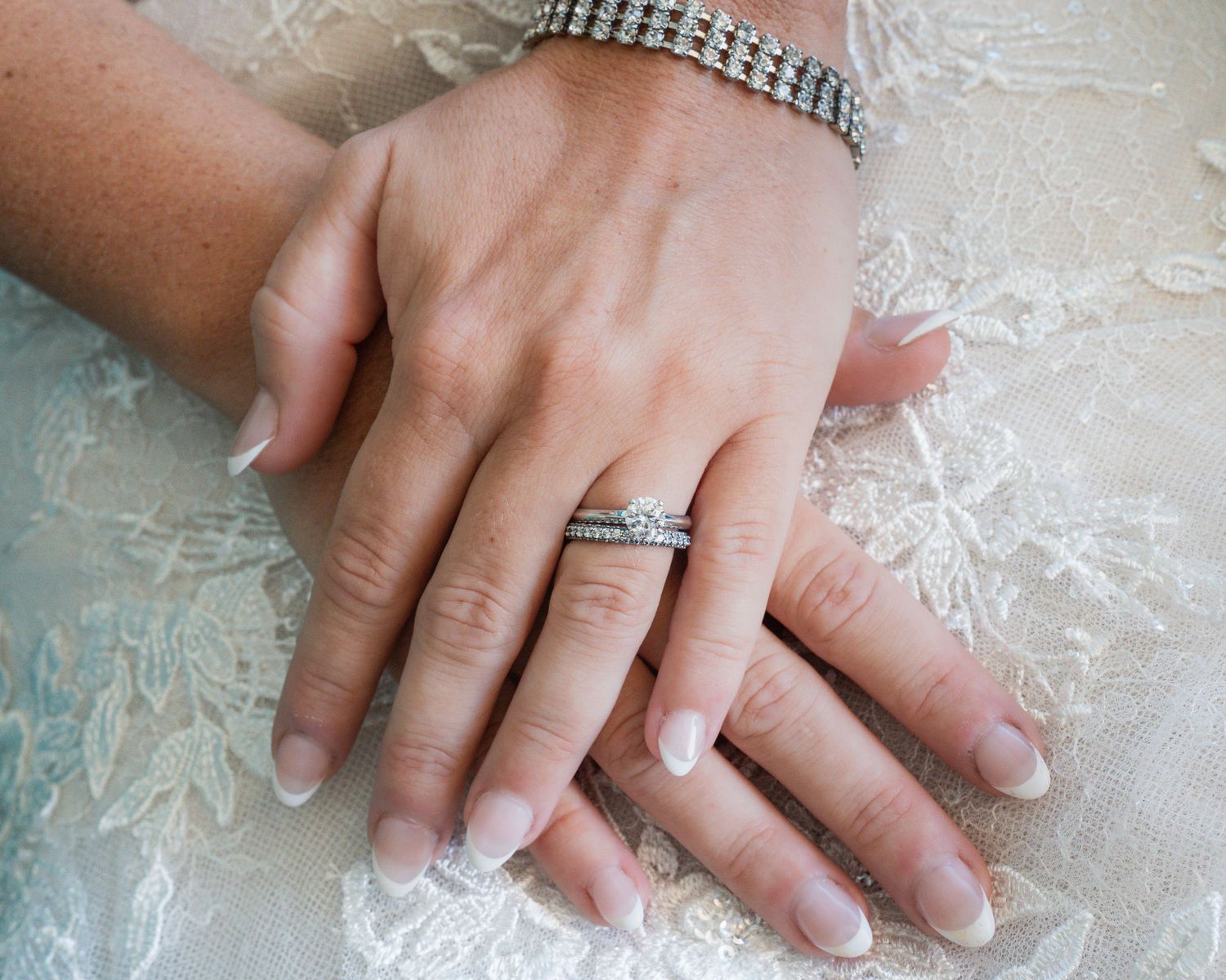 Close-up of bride and groom hands showing diamond wedding rings and sparkling tennis bracelet on white lace dress.