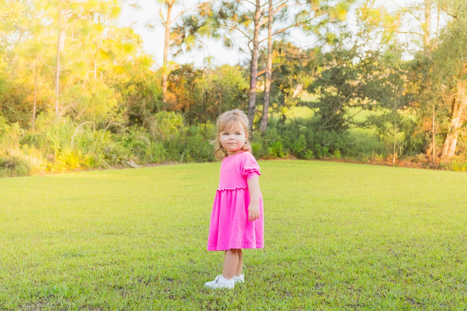 A toddler girl in a pink dress and white shoes stands on green grass with trees in the background.
