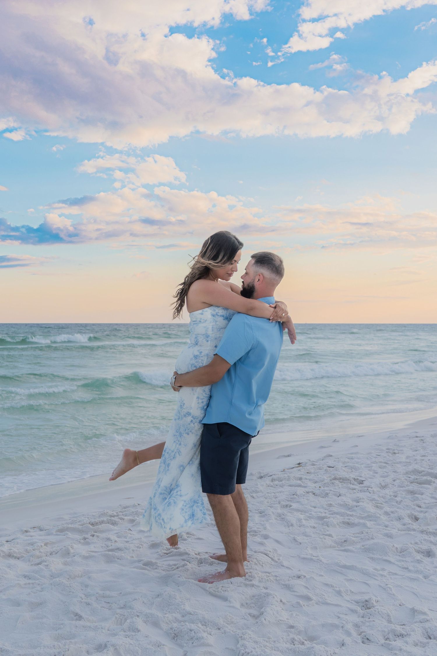 Couple embracing on a white sand beach at sunset, with pastel clouds and turquoise ocean waves in the background.