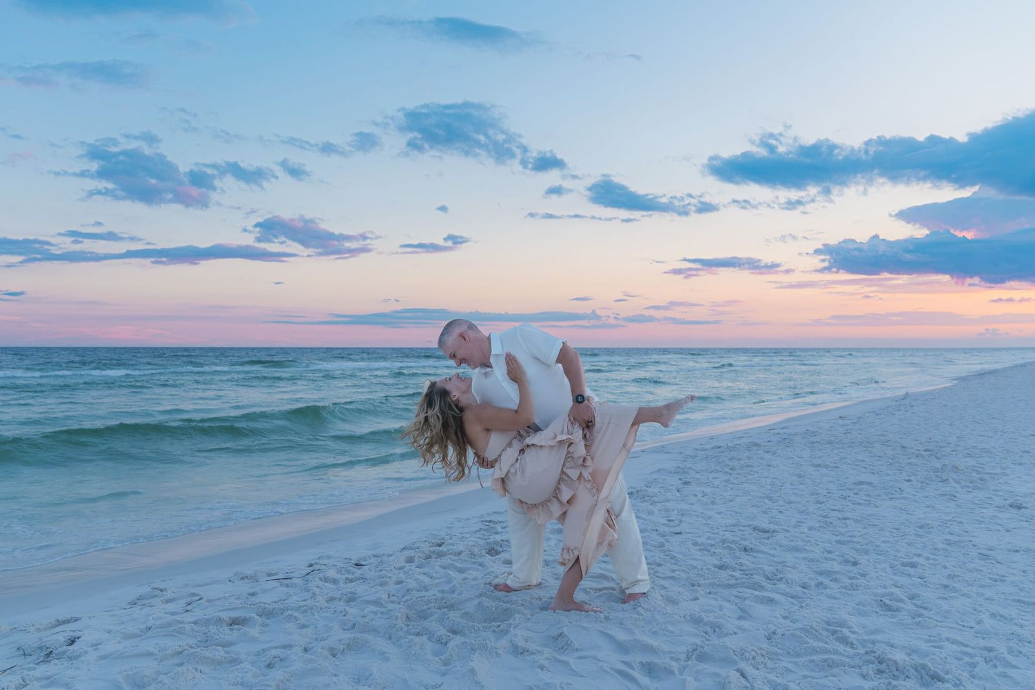 Couple dancing romantically on a white sand beach at sunset with pastel pink and blue skies.