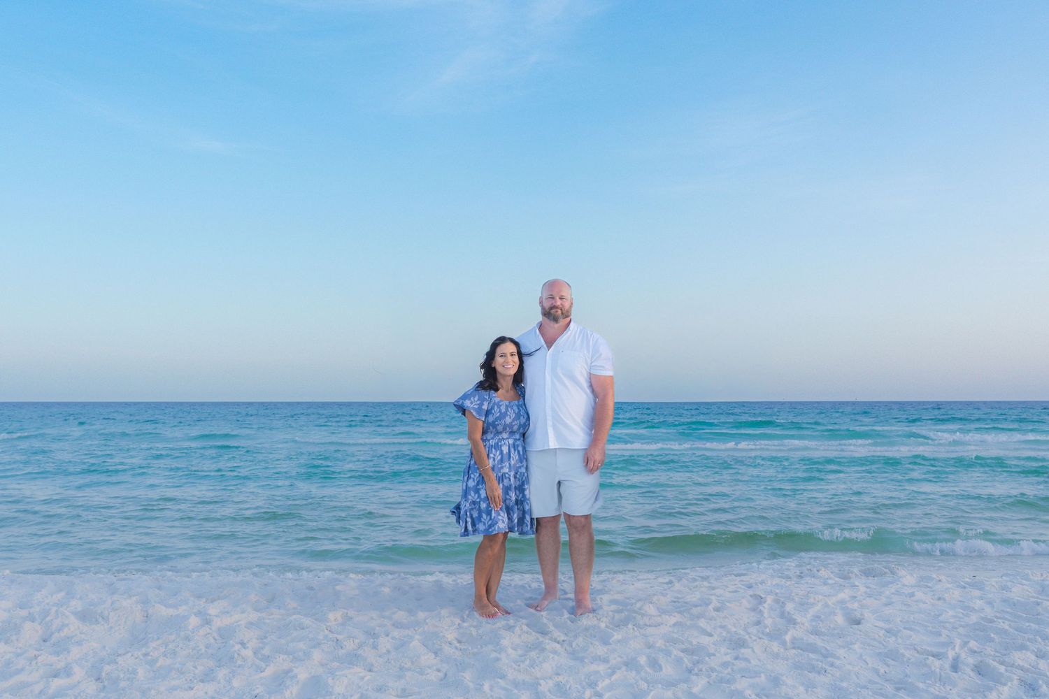 Couple standing on white sandy beach with turquoise ocean water and blue sky backdrop.