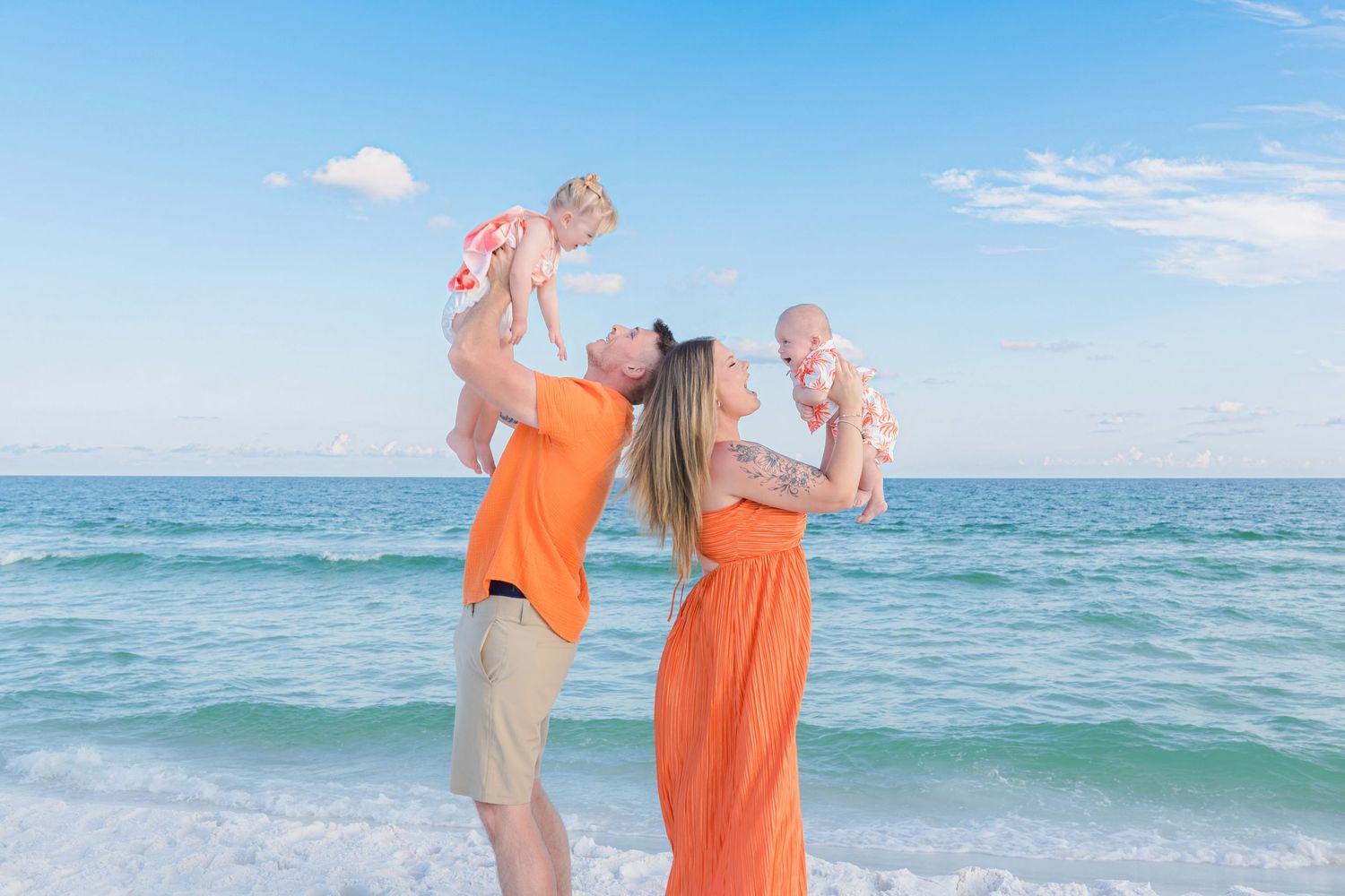 Family in matching orange outfits lifting babies at the beach with blue ocean and sky backdrop.