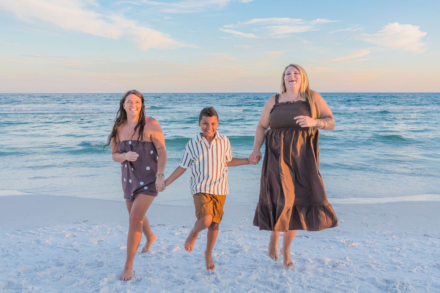 Family of three joyfully jumping on a white sand beach at sunset with turquoise ocean waves in the background.
