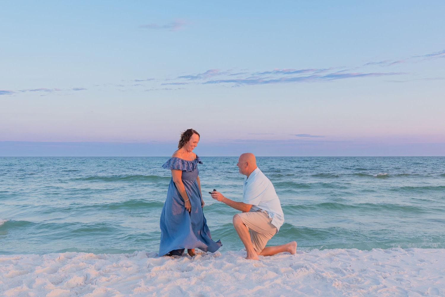 Man proposes to woman on a sandy beach at sunset with ocean waves in the background.