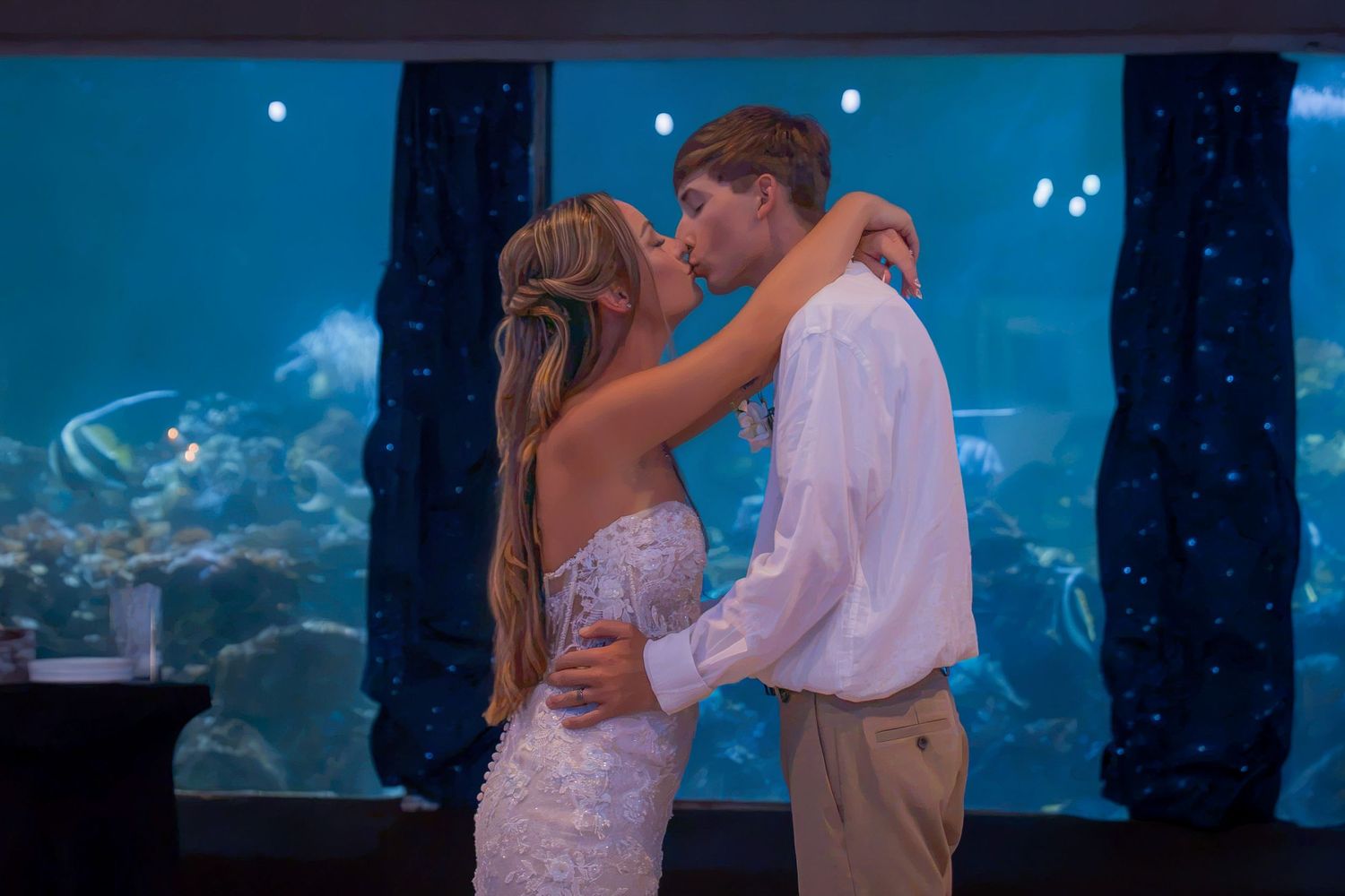 Couple sharing a romantic kiss in wedding attire in front of a large aquarium tank with tropical fish.