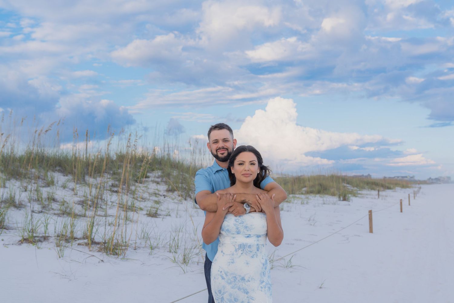 Couple embracing on white sand beach with sea oats and dramatic cloudy sky at sunset.