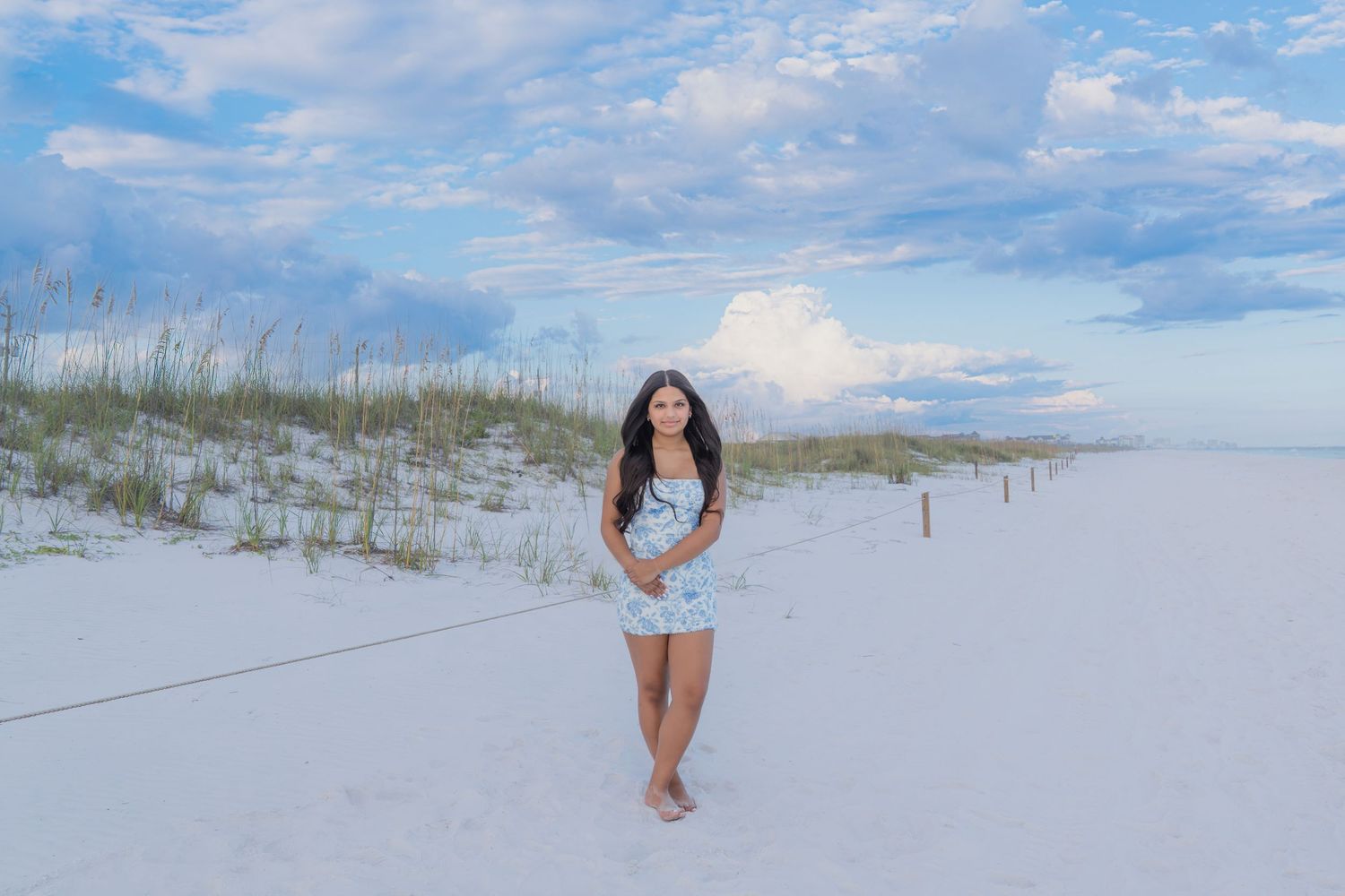 A young woman in a floral dress stands on white sand beach dunes under a dramatic cloudy blue sky.