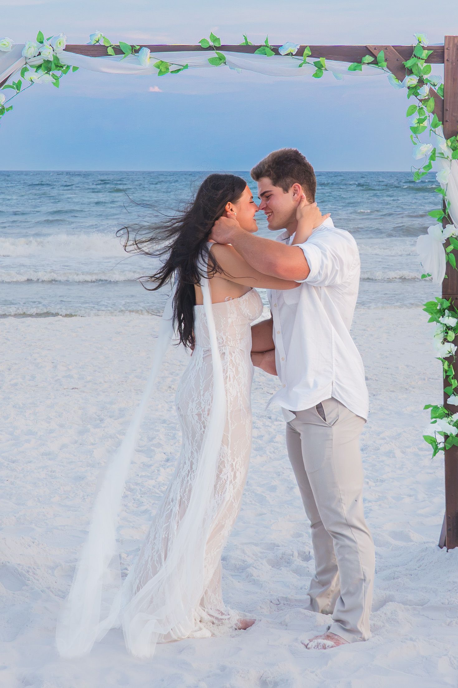 Couple embracing during a romantic beach wedding ceremony under a floral arch at sunset.