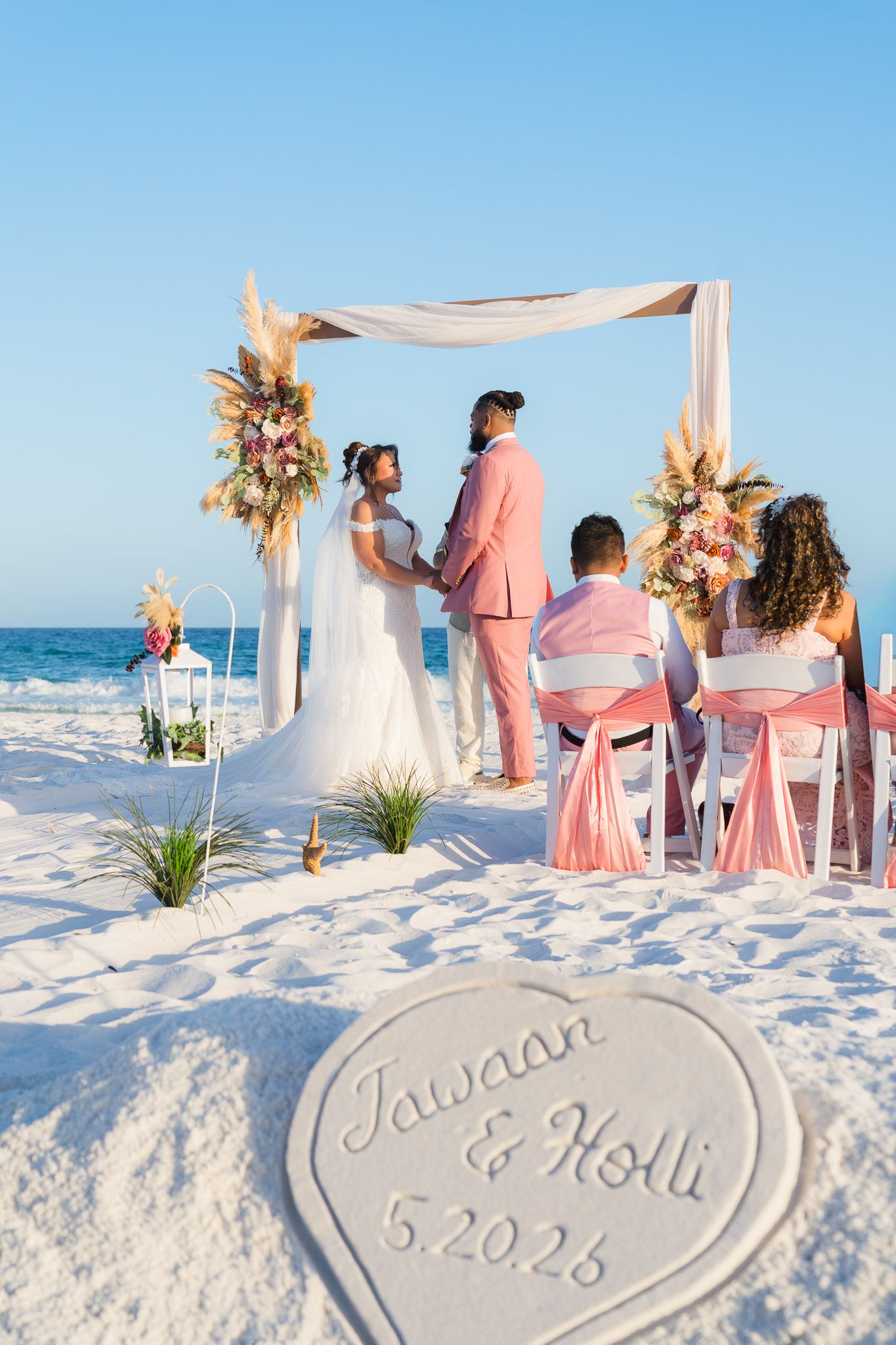 Beach wedding ceremony of Jawaan and Holli on 5.20.26, with couple at floral arch, guests seated in pink chairs on white sand.